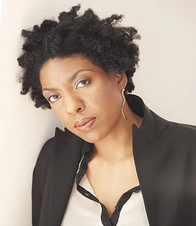 woman dressed in dark blazer and with a beige shirt and black trimming underneath.  She looks directly into camera. 