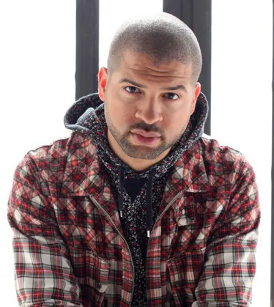 A man dressed in a red, white and black lumberjack jacket and tweed colored hood shirt stares directly into camera. 