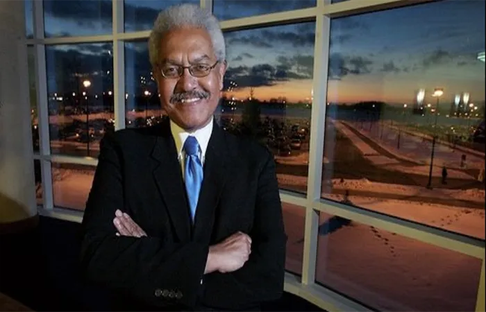 Dr. Manning Marable smiles with folded arms, dark suit, blue tie and white collared shirt in front of a picturesque window.