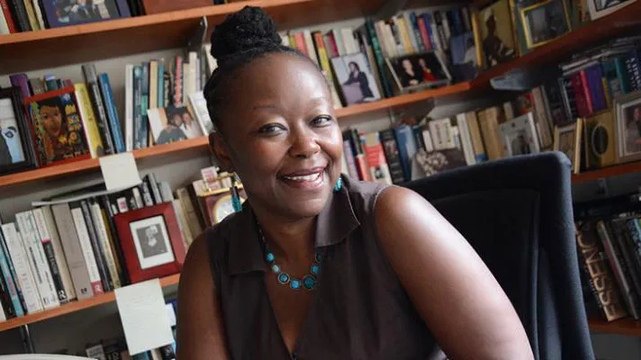 Farah Jasmine Griffin, wearing a sleeveless brown blouse and upswept braids, sits behind her office desk, book-lined shelves in the background