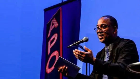 Architectural historian John Reddick lectures in front of a microphone against a backdrop showing an image of the Apollo Theatre in New York City