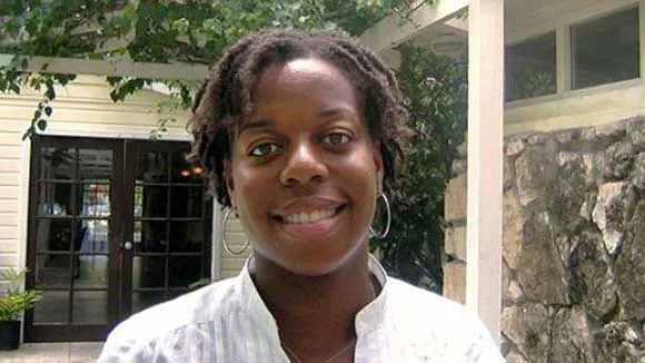 Historian Natasha Lightfoot, wearing a collarless white blouse and a chain with a heart dangling from it, smiles into the camera while standing in front of a brick facade on a sunny day. There is a small tree in the background.