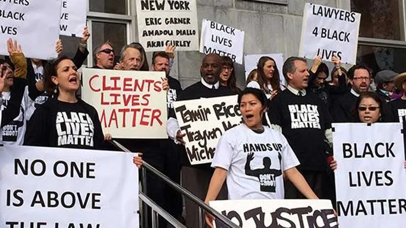 A group of roughly two dozen people stands on steps shouting and holding signs that read: "Black Lives Matter," "No Justice No Peace," "Lawyers 4 Black Lives Matter," "Client Lives Matter," and others.