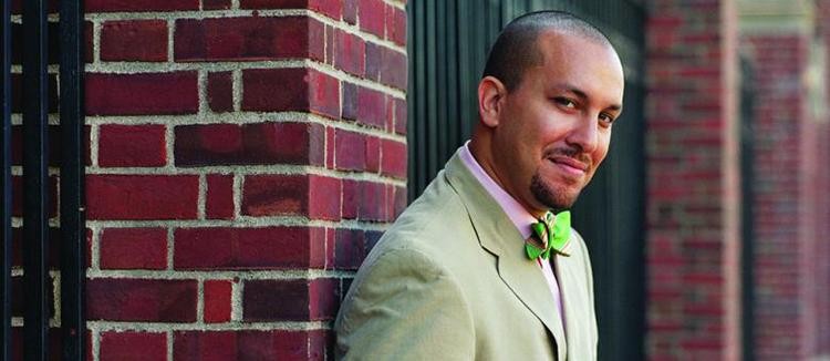 Daylight: Columbia University Religion Professor Josef Sorett leans against a red brick pillar, smiling slightly and wearing a pale green suit, pink button-down shirt, and bright green bow tie; photo by Gabriel Cooney.